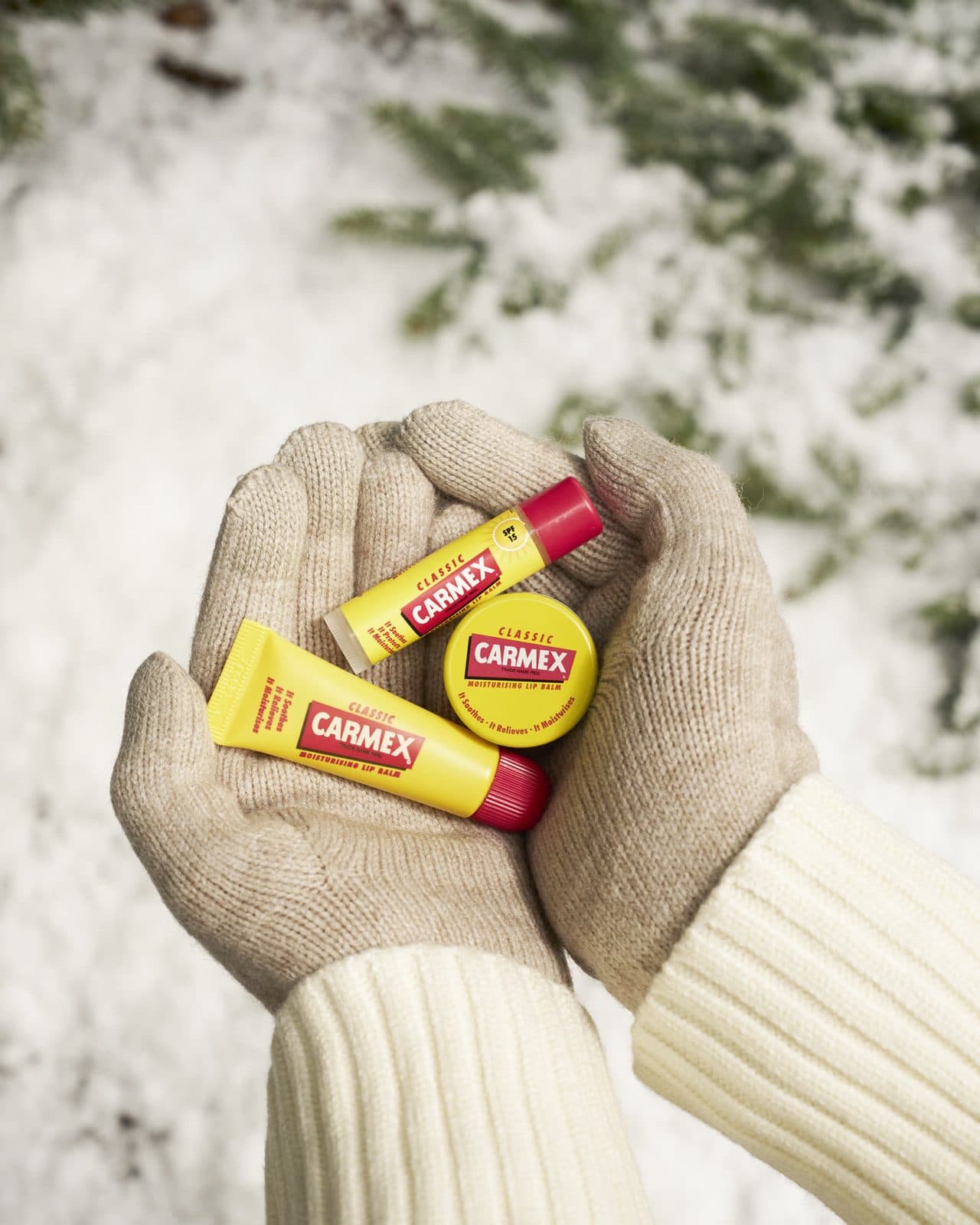 Kesztyűs kezek Carmex Classic tégelyt, tubust és stiftet tartanak a hó fölött